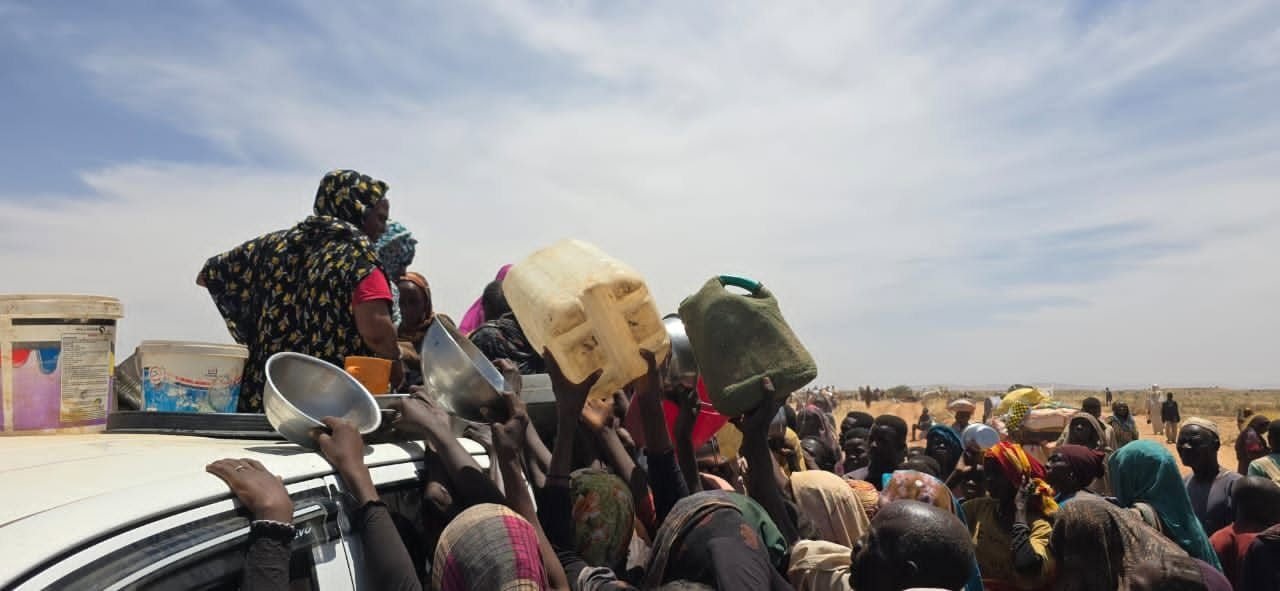 Displaced people reach up with bowls and water jugs to a relief vehicle in Tawila
