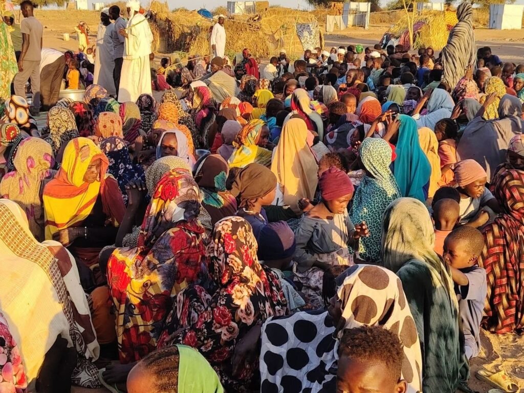 Families seated on the sand waiting for a meal, North Darfur, June 2025